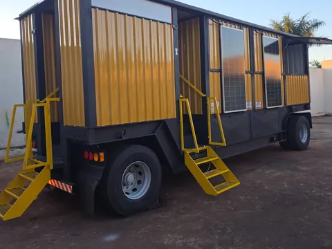 Trailer amarelo com toldo, escadas e caixa d'água, em três quartos contra o céu claro